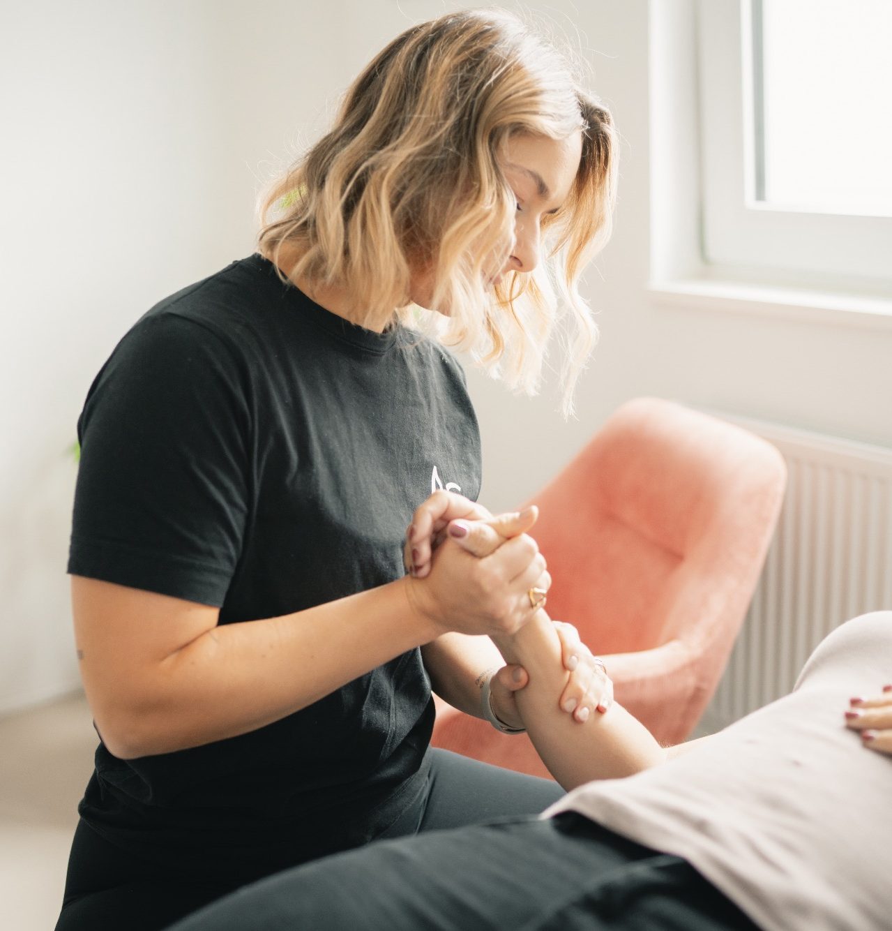 Frau massiert die Hand einer Person auf einer Liege in einem hellen Raum.